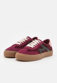 Paire de baskets en daim bordeaux avec lacets blancs, détails latéraux noirs et semelles en caoutchouc marron clair sur fond blanc.