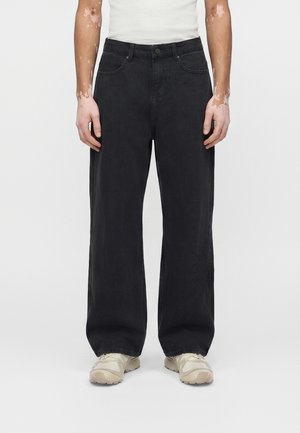 Άτομο που φοράει μαύρο τζιν με φαρδιά πόδια, λευκά αθλητικά παπούτσια και λευκό φανελάκι, στέκεται μπροστά σε απλό φόντο.