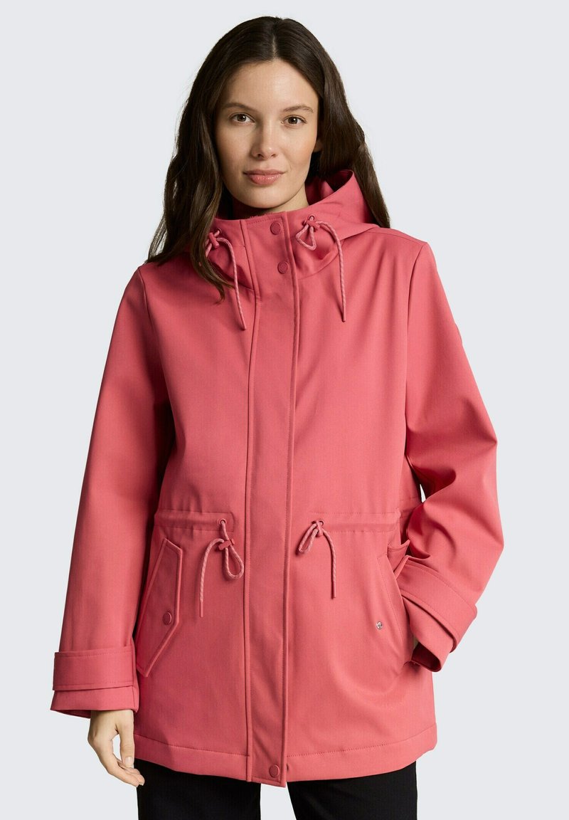 Frau trägt eine langärmelige korallenrosa Kapuzenjacke mit Kordelzügen und Taschen vorne, steht vor einem hellen Hintergrund.