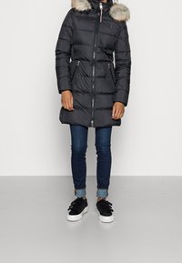 Osoba oblečená v černém prošívaném zimním kabátu s kožešinovou kapucí, modrých zkrácených džínech a černých teniskách s bílou podrážkou.