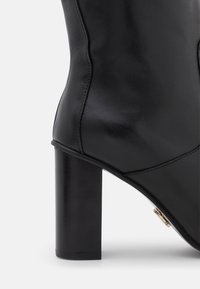 Czarny skórzany botek sięgający do kostki, z okrągłym noskiem, gładką fakturą i masywnym czarnym obcasem. Złote detale przy szwie bocznym.