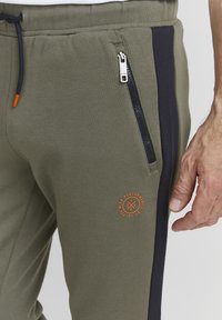 Olive green joggers with a black side panel, featuring a zippered pocket and an orange logo detail. Made of soft, stretchy fabric.