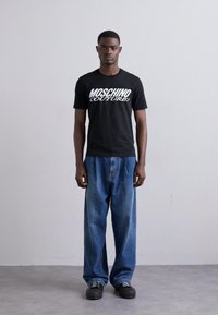 T-shirt nera in cotone con logo bianco "Moschino Couture!", abbinata a jeans in denim blu sbiaditi a vestibilità ampia e sneakers nere.