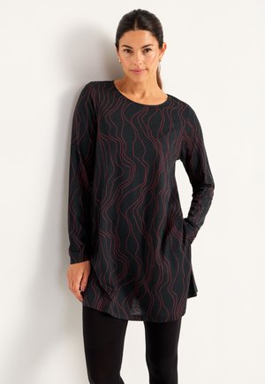 Vrouw met zwart haar draagt een lang mouw zwart tuniek met rode golvende lijnen, gecombineerd met zwarte leggings, terwijl ze tegen een witte muur staat.
