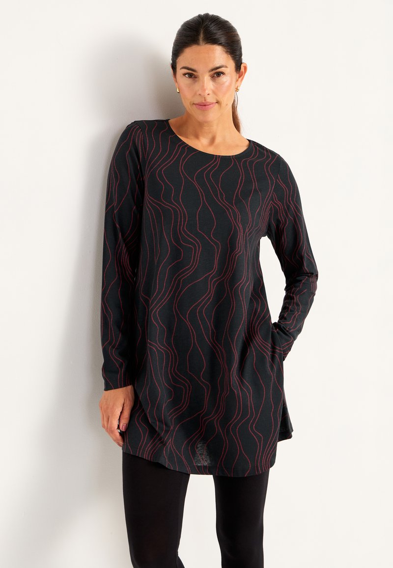 Kvinna med mörkt hår, klädd i en långärmad svart tunika med röda svängande linjer, i kombination med svarta leggings, står mot en vit vägg.