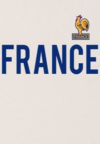 Bomull t-shirt i krämfärg med stor marinblå text som säger "FRANKRIKE." Har ett gult höns-emblem ovanför texten. Enkelt design, avslappnad passform.