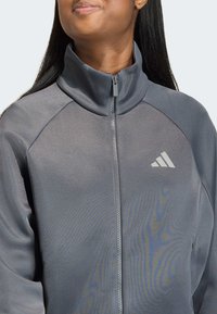 Γκρι αθλητικό μπουφάν με ψηλό γιακά, πλήρες φερμουάρ και μαλακή υφή· διαθέτει διακριτικό λογότυπο και λεπτομέρειες ραφών στους ώμους.