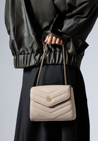 Borsa a tracolla beige trapuntata con una tracolla a catena, con logo dorato e motivo a zigzag. Abbinata a una giacca di pelle scura e una gonna nera.