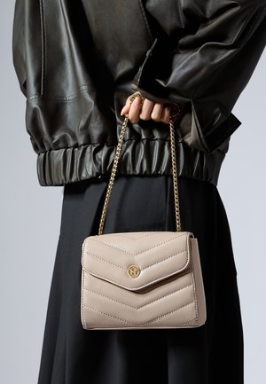 Sac à main beige matelassé avec une bandoulière en chaîne, arborant un logo doré et un motif en chevrons. Associé à une veste en cuir noire et une jupe noire.
