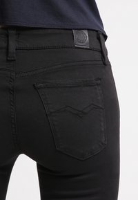 Jeans de mezclilla negra con un diseño ajustado, que presentan un patrón cosido en los bolsillos traseros y un parche de cuero con logo en la cintura.