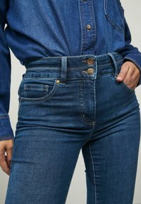 Temno modri kavbojke z visokim pasom, ki imajo dva sprednja žepa, dvojno zapiranje s gumbi in gladko teksturo.