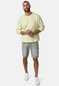 Felpa gialla chiaro con scollo a girocollo, abbinata a pantaloni corti in denim grigi con risvolti e un accento verde. Sneakers bianche completano il look.