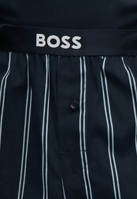 Μαύρα boxer με κάθετες λευκές ρίγες και κουμπιά στο μπροστινό μέρος, με μαύρη μέση που φέρει τη λέξη «BOSS» με λευκά γράμματα.