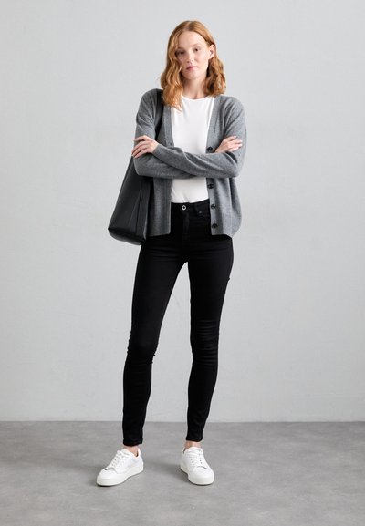 Grauer Cardigan mit Knopfdetails, getragen über einem weißen Hemd, kombiniert mit schwarzen Skinny Jeans und weißen Sneakers, vor einem hellgrauen Hintergrund.