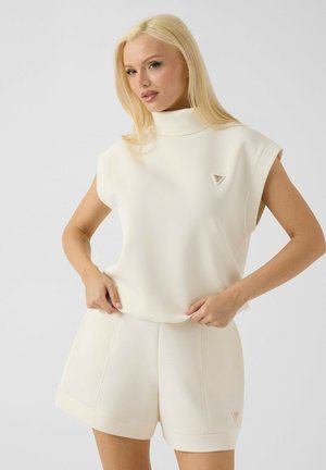 Donna con lunghi capelli biondi che indossa un top bianco senza maniche a collo alto e shorts bianchi abbinati su uno sfondo chiaro uniforme.