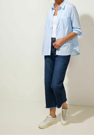 Chemise rayée bleu clair avec manches retroussées, deux poches avant, associée à un jean court bleu marine et des baskets blanches.