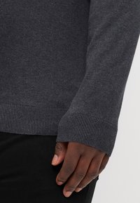Maglione grigio scuro con una consistenza morbida, maniche aderenti e polsini a costine, abbinato a pantaloni neri, con una mano appoggiata sul tessuto.