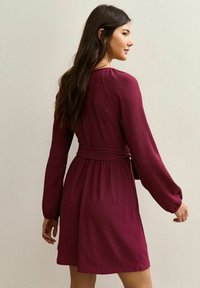 Bordowa sukienka z długimi rękawami i paskiem w talii, z teksturowanej tkaniny i spódnicą do kolan, stylizowana na dopasowaną sylwetkę.