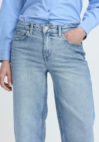 Šviesiai mėlynos džinsų kelnės su laikinos formos siluetu, turinčios priekinė kišenes, mygtuką ir subtilius išblukimus. Medžiaga atrodo lygi.
