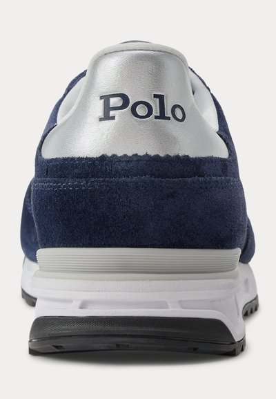 Achteraanzicht van een marineblauwe suède sneaker met zilveren hielstukje en witte zool, met de merknaam "Polo" in zwarte tekst.