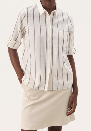 Personne portant une chemise blanche à manches longues boutonnée avec des rayures verticales noires et une jupe beige, une main dans la poche et les manches retroussées.