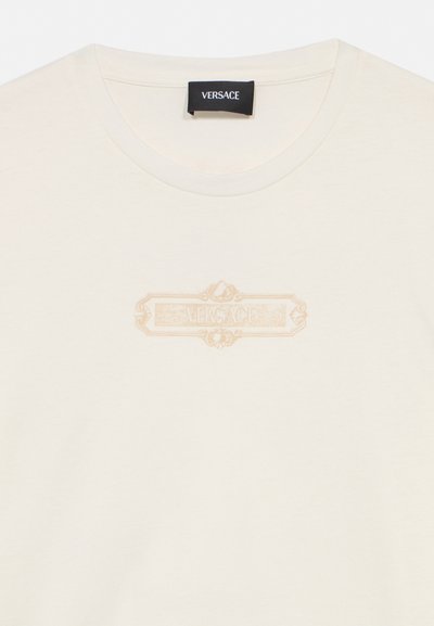 T-shirt Versace écru avec col rond et logo orné beige subtil centré sur la poitrine sous une étiquette de marque noire à l’intérieur du col.