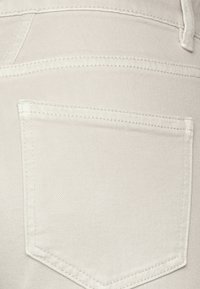 La tela de mezclilla de un beige claro presenta un bolsillo trasero con costuras dobles y una textura tejida, creando un aspecto limpio y estructurado.