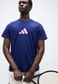 Αθλητική μπλούζα σε ναυτικό μπλε χρώμα, κατασκευασμένη από βαμβάκι, με μεγάλο ροζ λογότυπο Adidas και κείμενο "PADDEL" στον αριστερό ώμο. Στενή εφαρμογή.