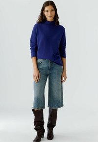 Paarse gebreide trui met een hoge hals en een zijzak, gecombineerd met vervaagde wijde denim jeans en bruine knielange laarzen.