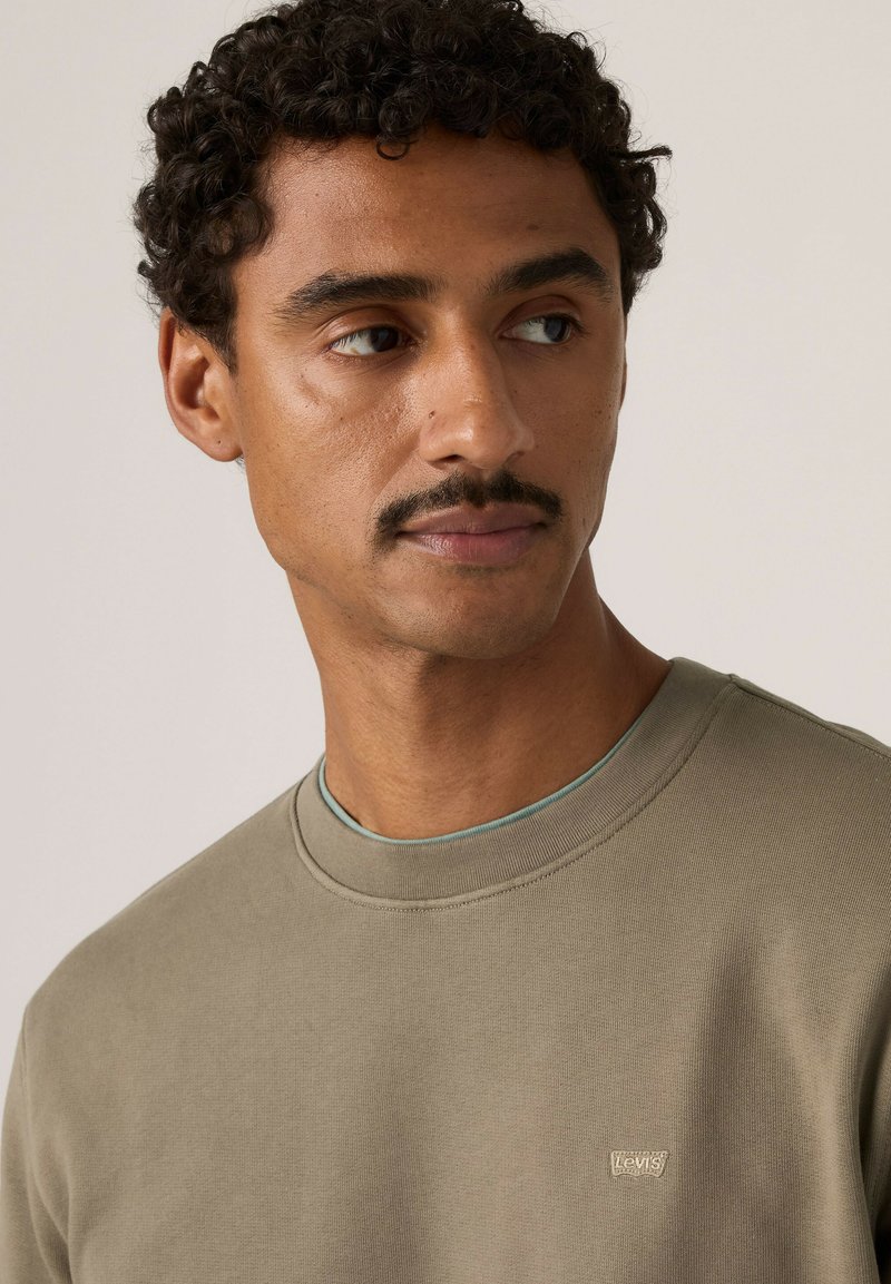 Mann mit kurzen lockigen Haaren und Schnurrbart, der einen olivgrünen Levi's Rundhalsausschnitt-Sweatshirt trägt und seitlich vor schlichtem Hintergrund blickt.