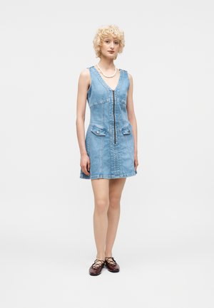 Γυναίκα με κοντά σγουρά ξανθά μαλλιά που φοράει αμάνικο ανοιχτό μπλε τζην φόρεμα με φερμουάρ μπροστά και καφέ δαντελωτά παπούτσια.