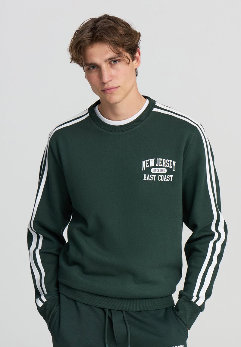 Felpa verde con righe bianche sulle maniche, collo tondo e testo "New Jersey East Coast" sul davanti. Tessuto liscio, vestibilità casual.