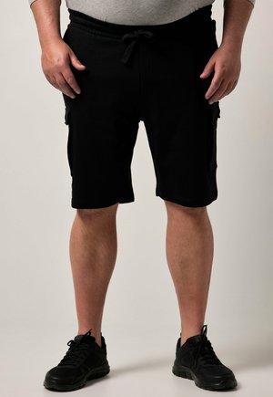Man draagt zwarte cargoshorts en zwarte sneakers, staat met ontspannen handen langs zijn zij tegen een effen achtergrond.