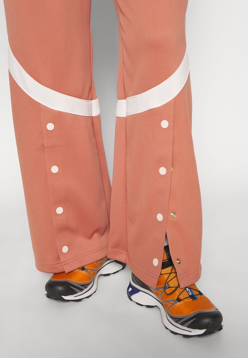 Pantalons de sport roses avec des accents blancs et des boutons-pression au bas, associés à des chaussures de course orange et noires.