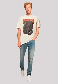 Beige Baumwoll-T-Shirt mit einem grafischen Design, das eine Schlange zeigt, kombiniert mit hellblauen, verwaschenen Jeans und weißen Sneakers.