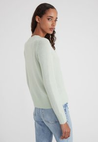 Frau mit lockigem Haar, die einen hellgrünen Pullover und blaue Jeans trägt und über die Schulter auf einen schlichten weißen Hintergrund blickt.