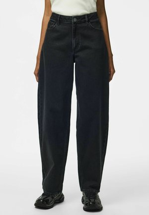 Person trägt locker sitzende schwarze Jeans und schwarze Sneaker, steht vor einem einfarbigen weißen Hintergrund.