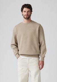 Beige sweatshirt met een ronde hals en ribgebreide manchetten, met een klein logo op de borst, gedragen over een wit shirt en lichte broek.