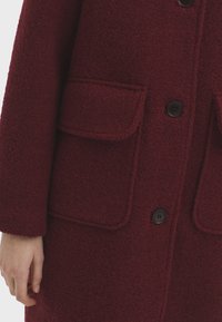 Bordeauxfarbener Mantel aus strukturiertem Stoff, mit großen Klappentaschen, einer Knopfleiste und einer lockeren Silhouette mit sichtbaren Nähdetails.