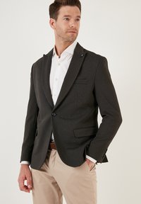Blazer grigio carbone in tessuto testurizzato, con un bottone e due bottoni, rever a punta e taschino sul petto. Indossato su una camicia bianca.