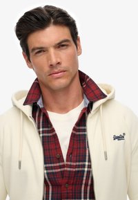 Hvid fleece hoodie lagdelt over en rød og marineblå ternet skjorte, med en denimkrave og mørke knapper, samt et logo på brystet.