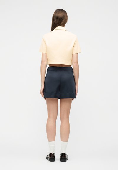 Γυναίκα με μακριά καστανά μαλλιά που φοράει κοντομάνικο μπεζ cropped μπλουζάκι, σκούρο μπλε σορτς, λευκές κάλτσες και μαύρα loafers, γυρισμένη πλάτη σε λευκό φόντο.