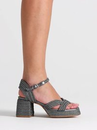 Jambe portant une sandale argentée métallique à talon haut avec talon bloc et boucle à la cheville sur un fond clair uni.