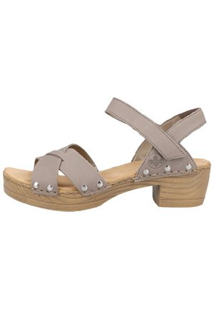 Sandale beige avec un petit talon bloc en bois, deux brides croisées à l'avant avec des clous métalliques et une bride de cheville réglable avec fermeture Velcro.
