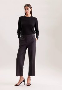 Maglione nero a manica lunga abbinato a pantaloni neri a vita alta e gamba larga. La modella indossa tacchi neri con cinturini. Tessuti lisci e setosi.