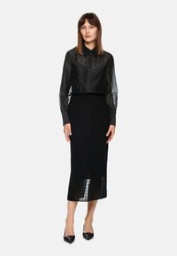Schwarze durchsichtige Bluse mit Knöpfen und langen Ärmeln, kombiniert mit einem figurbetonten schwarzen Midirock mit einem strukturierten Punktmuster. Schwarze spitz zulaufende High Heels.
