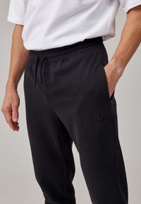 Zwarte joggingsbroek met een elastische tailleband en zijzakken, voorzien van een subtiel logo. Het materiaal lijkt zacht en textuur te hebben.