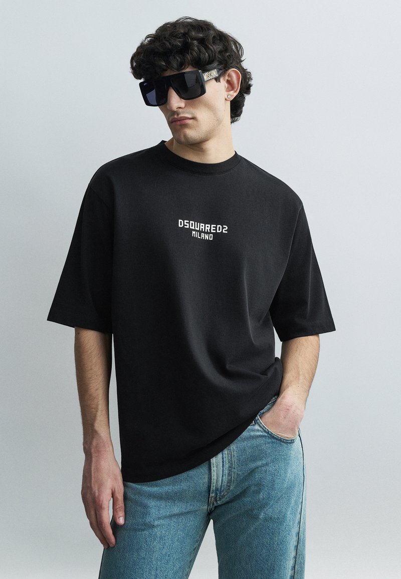 Melna kokvilnas T-krekls ar īsām piedurknēm un DSQUARED2 Milano logotipu baltā krāsā. Valkāts kopā ar gaiši zilām džinsa biksēm. Lielas melnas saulesbrilles.