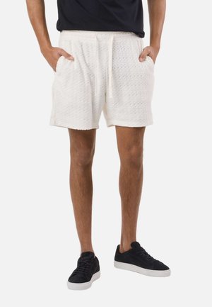 Mężczyzna w białych, teksturowanych szortach ze ściągaczem, czarnych tenisówkach z białymi podeszwami, stoi z rękami w kieszeniach.