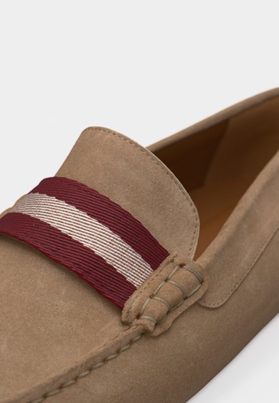 Mocassin en daim beige avec détails de couture et une bande en tissu rayée bordeaux et blanche sur la partie supérieure.